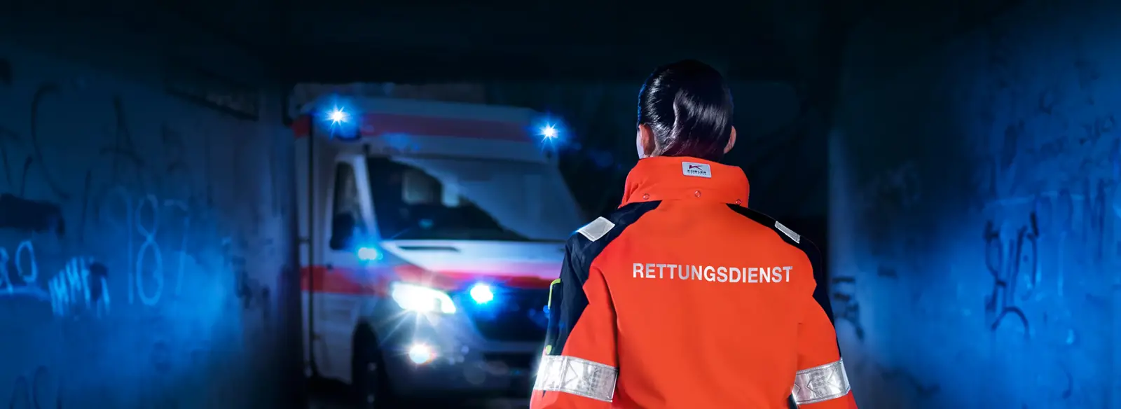 Eine Rettungskraft trägt auffällige, nach DIN EN ISO 20471 genormte Warnschutzkleidung von DBL und steht vor einem Rettungswagen. Die Kleidung bietet Sicherheit und Komfort bei jeder Witterung und wird im DBL Mietservice hygienisch aufbereitet und direkt in die Spinde einsortiert. Ideal für den anspruchsvollen Arbeitsalltag im Rettungsdienst.