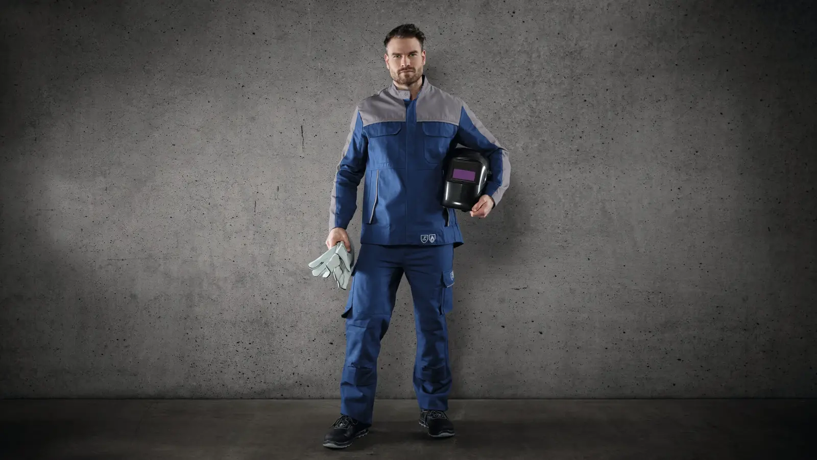 Ein Schweißer steht in moderner Schweißerschutzkleidung mit Helm und Handschuhen vor einer grauen Wand. Die Schutzkleidung entspricht den Normen EN 11611 und EN 11612 und bietet Einsätze aus Stretchgewebe für mehr Bewegungsfreiheit.